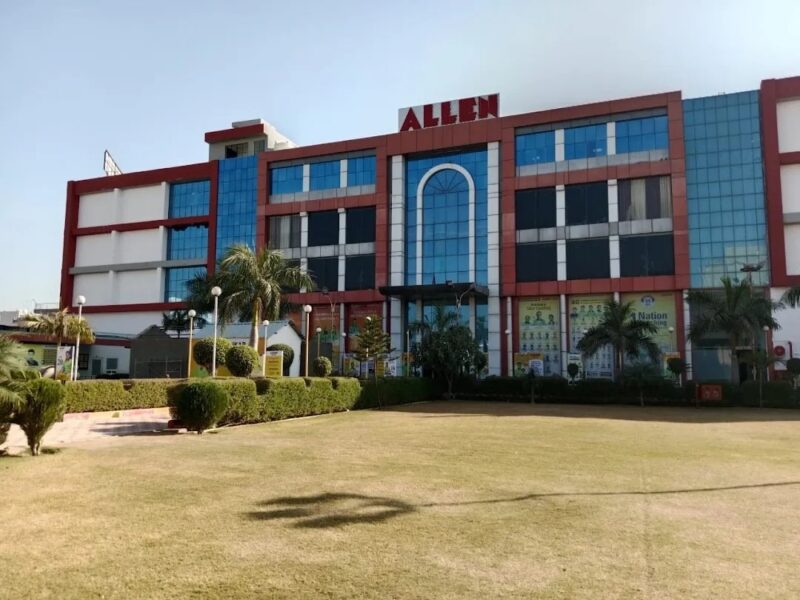 ALLEN Career Institute building in Sikar, Rajasthan. A large modern multi-story campus with red and blue glass facade, prominent "ALLEN" signage on the rooftop, well-maintained green lawn, palm trees, and landscaping in front.
